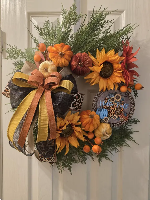 An autumn wreath on a white door, with sunflowers, orange pumpkins, cedar and leopard-print ribbon.